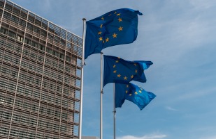 3 European Union flags flying in the wind next to a tall city building.